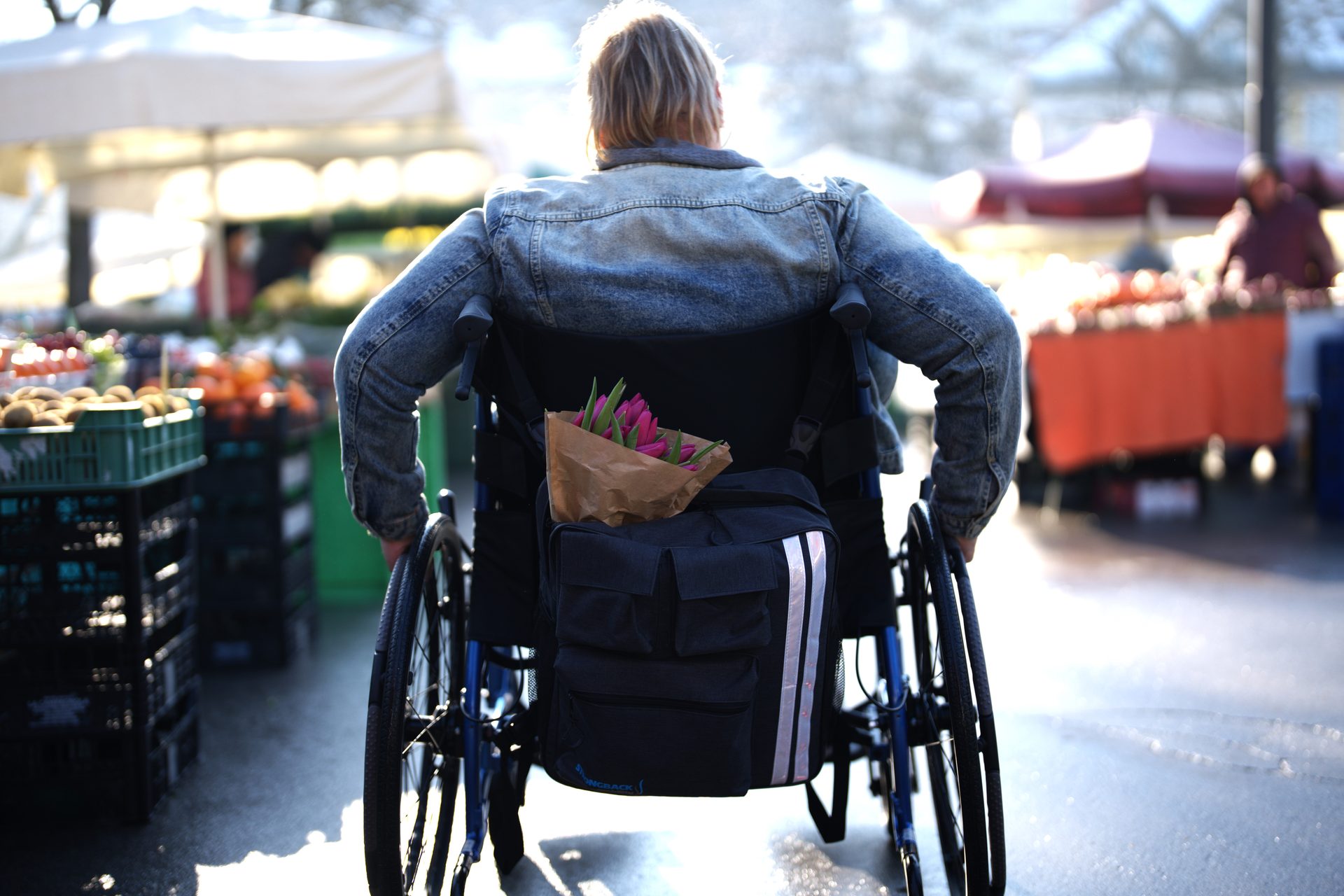 Person im Rollstuhl mit Rucksack und Blumenstrauß auf einem Wochenmarkt